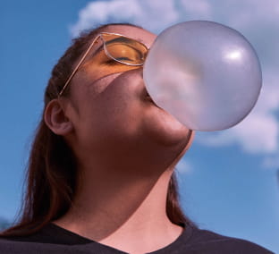 A imagem mostra uma mulher jovem, branca, de cabelos lisos, compridos e ruivos. Ela está usando camiseta preta e óculos de sol amarelos. Sob um céu azul de fundo, ela está de olhos fechados, com a cabeça virada para o céu, enquanto faz uma bola de chiclete.